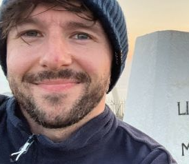 Chris is standing outdoors on a rocky hilltop at sunset. In the foreground, there is a white stone monument with the engraved words “LIVE FOR THE MOMENT.” A circular stone structure is visible to the right, surrounded by a decorative patterned platform. The landscape stretches out toward the coastline below, with a glowing orange sun setting over the distant water and casting warm light across the sky.